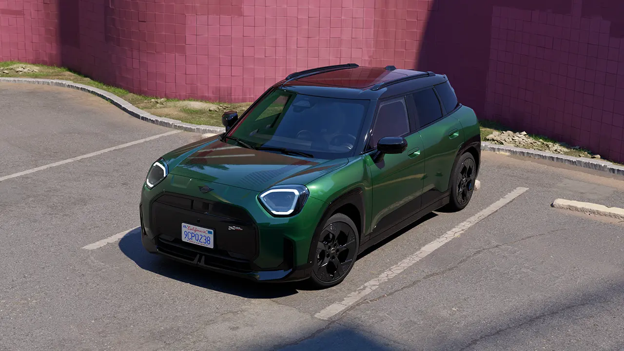 Imagine din față la parcarea unui MINI Aceman electric de culoare British Racing Green, într-o parcare cu un zid roz pe fundal