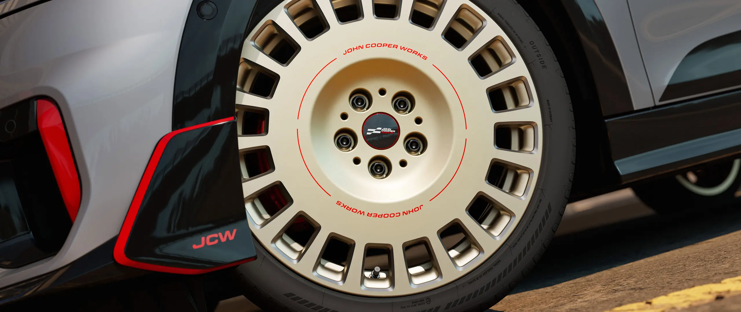 Prim-plan cu detaliile unei jante de 18" JCW Rallye, tip 964, spiţate, de culoare Vibrant Silver şi cu finisaj diamantat, alături de spoiler faţă.