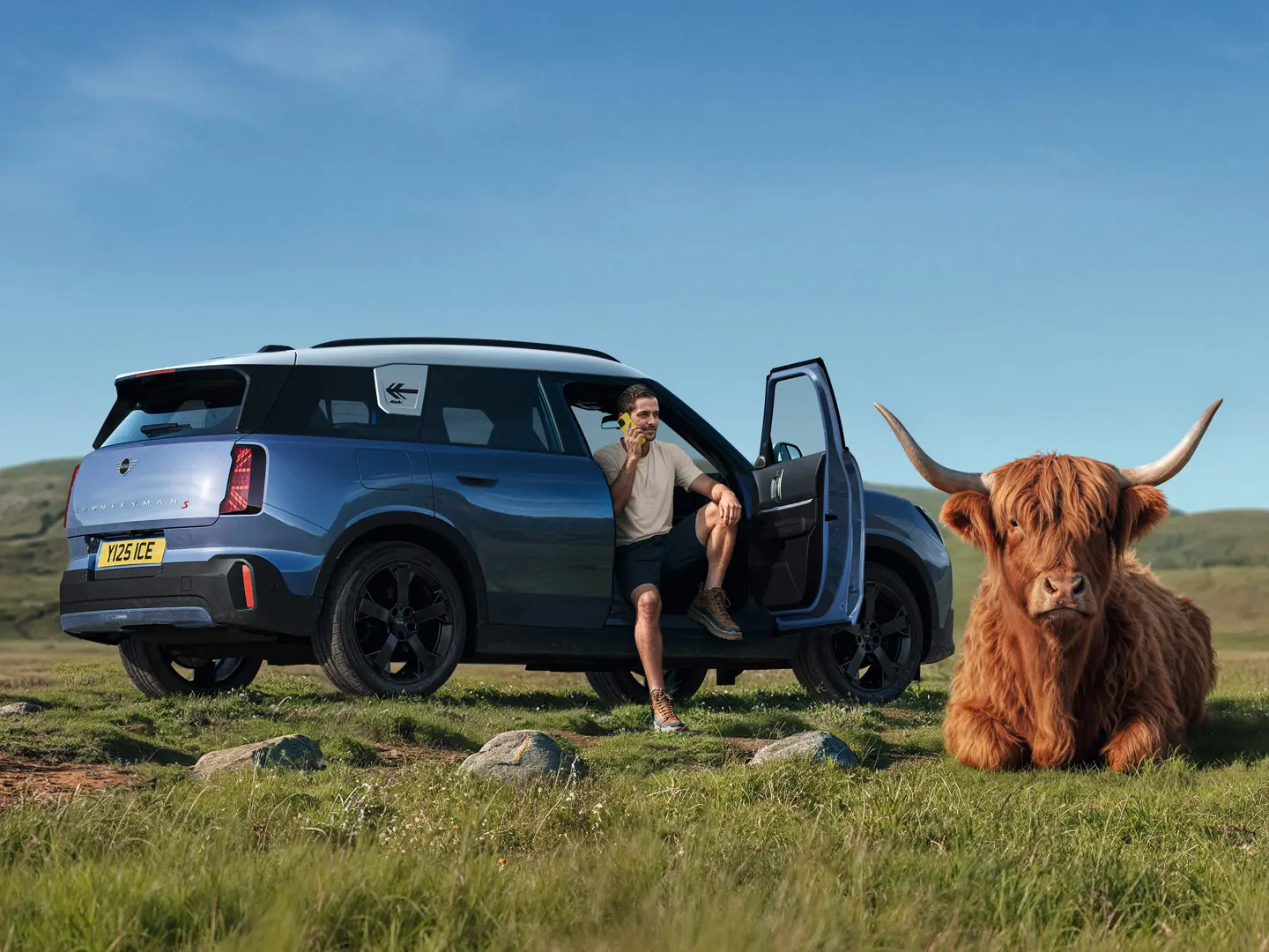 Un bărbat în fața automobilului său MINI Countryman S, vorbind cu serviciul de Asistență rutieră MINI prin intermediul aplicației MINI, și un taur Highland întins lângă el.